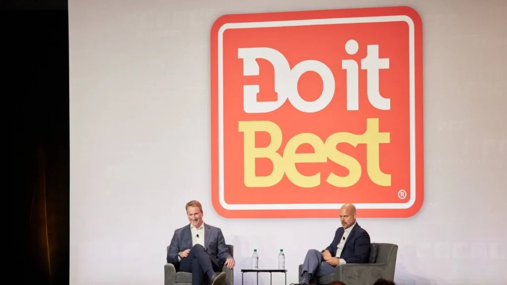 Two men sitting on stage at a Do It Best corporate event, engaging in discussion beside a large Do It Best logo